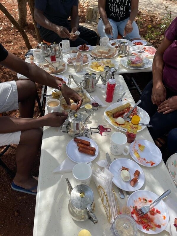 Un groupe de personnes est assis autour d’une table, devant divers plats et boissons, notamment des saucisses, des œufs et du café.