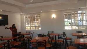 Um restaurante com várias mesas e cadeiras, uma televisão na parede e janelas grandes com painéis de vidro decorativos.