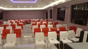 Sala de conferências com fileiras de cadeiras brancas e vermelhas, grandes janelas e luminárias no teto.