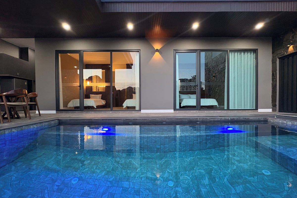 Ein moderner Poolbereich mit klarem Pool, überdachter Sitzgruppe und einem durch Glastüren einsehbaren Schlafzimmer.