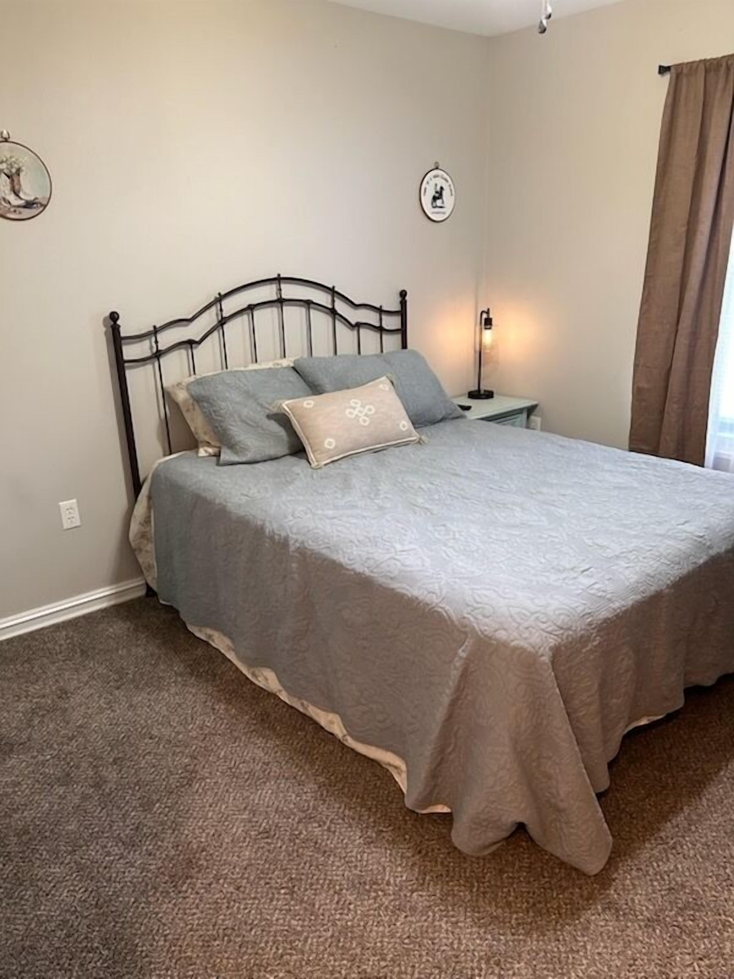 A neatly made bed with a grey bedspread and pillows, a bedside table with a lamp, and a wall-mounted picture frame.
