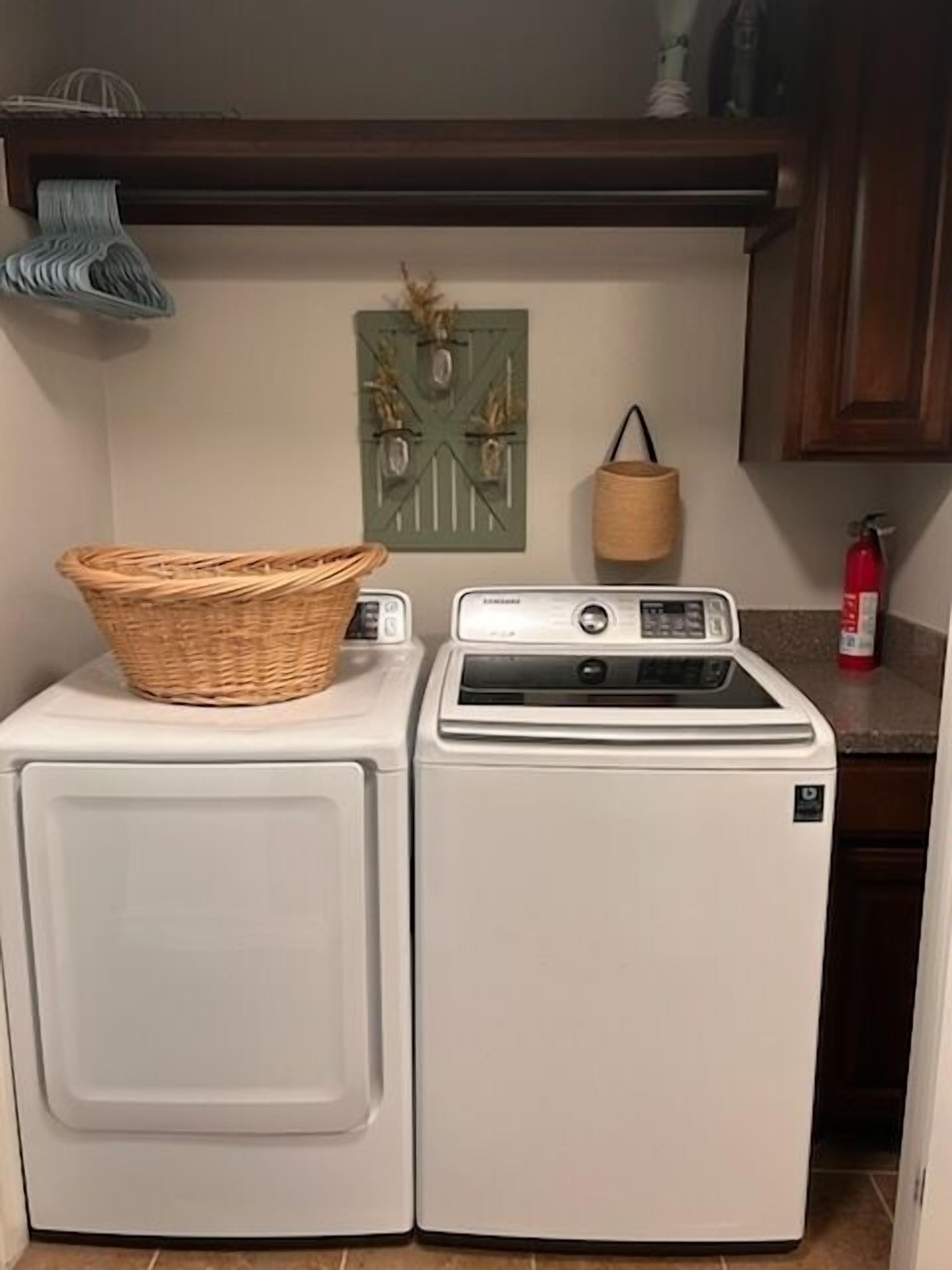 A compact laundry area with a front-loading washer and dryer, a wicker basket on the dryer, a wall-mounted basket holder, and a fire extinguisher on the countertop.