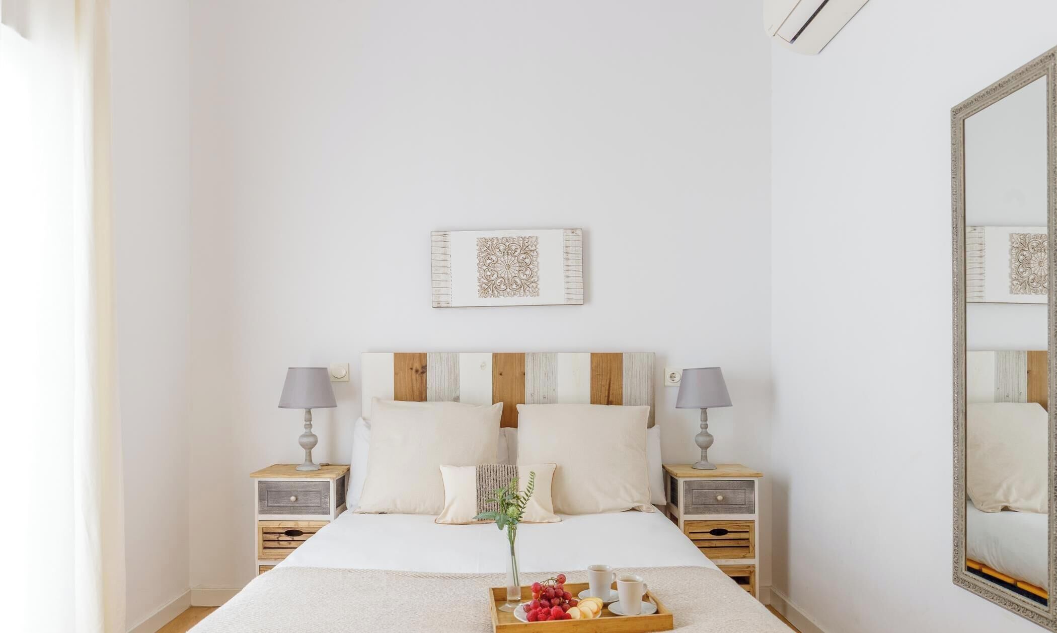 Un dormitorio ordenado con una cama grande, dos mesitas de noche con lámparas, una bandeja de fruta y café, y un espejo en la pared.