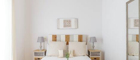 Un dormitorio ordenado con una cama grande, dos mesitas de noche con lámparas, una bandeja de fruta y café, y un espejo en la pared.