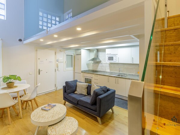 Un salon moderne avec un canapé, une table basse et des fauteuils, une cuisine avec des meubles blancs et des appareils électroménagers, et un escalier doté de rampes en verre.