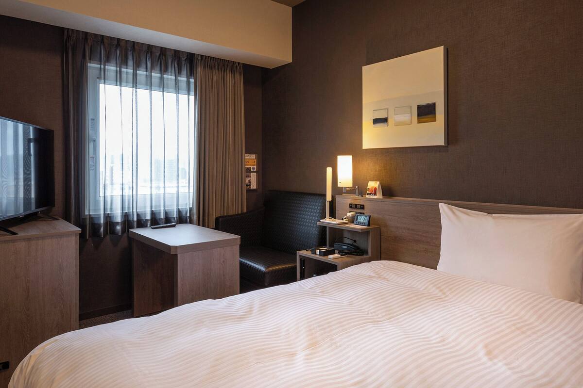 Ein Hotelzimmer mit Bett, Schreibtisch, Stuhl, Fernseher und einem Fenster mit Vorhängen.