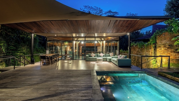 A modern outdoor pool area with a wooden deck, glass-enclosed lounge, and a covered seating area. - Twin House Ubud by Betterplace (Mas)