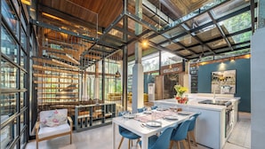 A modern kitchen with a central island, a dining area with blue chairs, and a staircase with a glass railing. - Twin House Ubud by Betterplace (Mas)