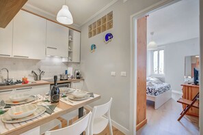 Una cocina moderna con armarios blancos, una mesa de comedor lista para cuatro personas y vistas a un dormitorio con cama y televisor.