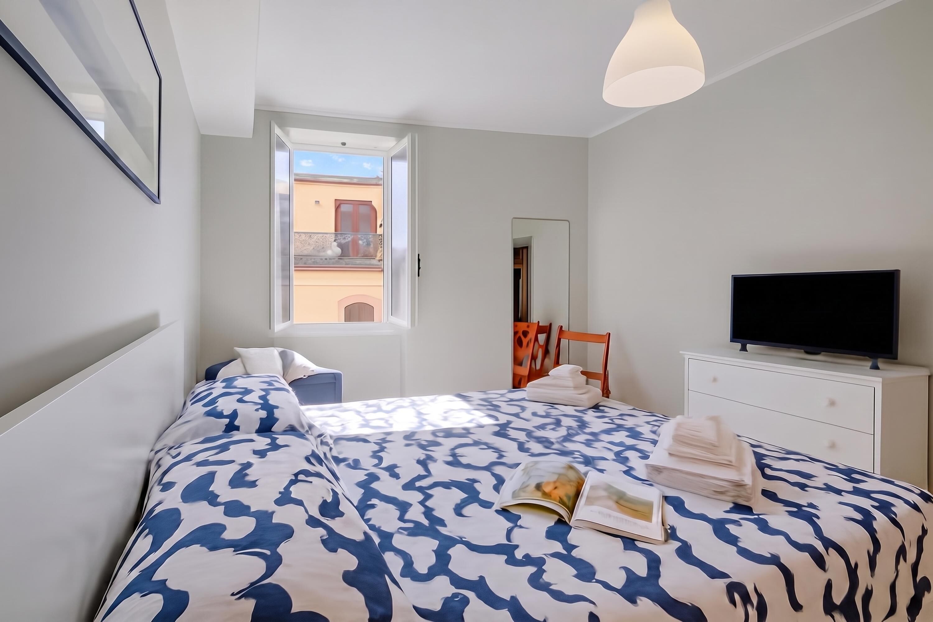Un dormitorio con cama, televisor, cómoda y ventana con vistas a edificios.