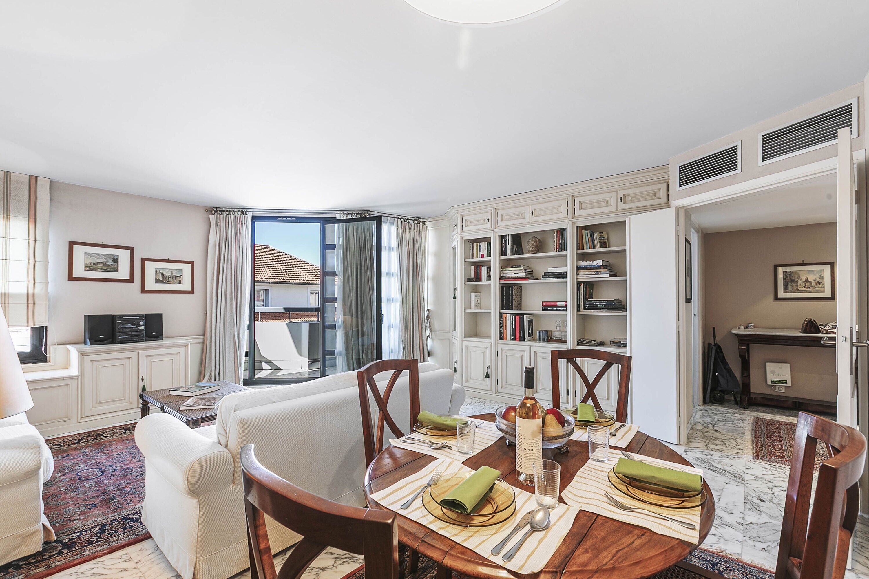 A dining area with a round table set for four, surrounded by white armchairs. A bookshelf filled with books and decorative items is visible in the background.