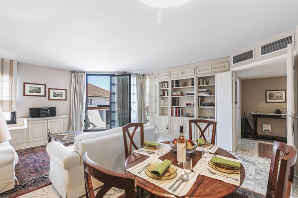 A dining area with a round table set for four, surrounded by white armchairs. A bookshelf filled with books and decorative items is visible in the background.