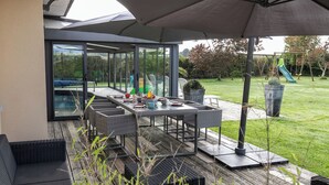 An outdoor patio with a dining table and chairs, surrounded by glass doors and a view of a garden and playground. - Chez David by Interhome (Kersaint-Plabennec)