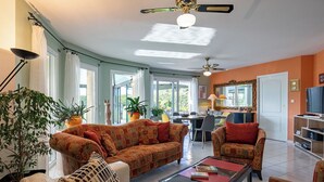 A spacious living room with a large sofa set, a coffee table, and a dining area with a table and chairs. There are multiple potted plants and a ceiling fan. - Chez David by Interhome (Kersaint-Plabennec)