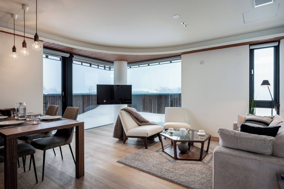 A modern living room with a wooden dining table, comfortable seating, a flat-screen TV, and large windows offering a view of the outdoors.