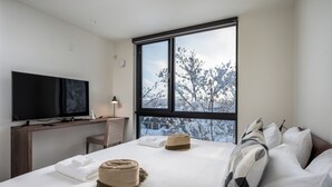 A modern hotel room with a large window, a flat-screen TV, a desk, and a neatly made bed with towels and a hat. - Yotei Dream One (Kutchan)