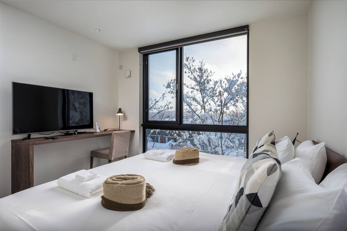 A modern hotel room with a large window, a flat-screen TV, a desk, and a neatly made bed with towels and a hat.