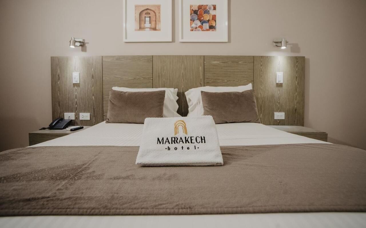 A hotel room with a wooden headboard, a bed with brown bedding, a white towel with the hotel name, and two framed pictures on the wall.