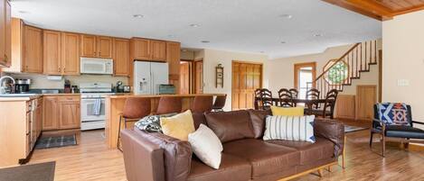 A spacious living area with a brown leather sofa, wooden cabinets, a kitchen area, and a staircase.