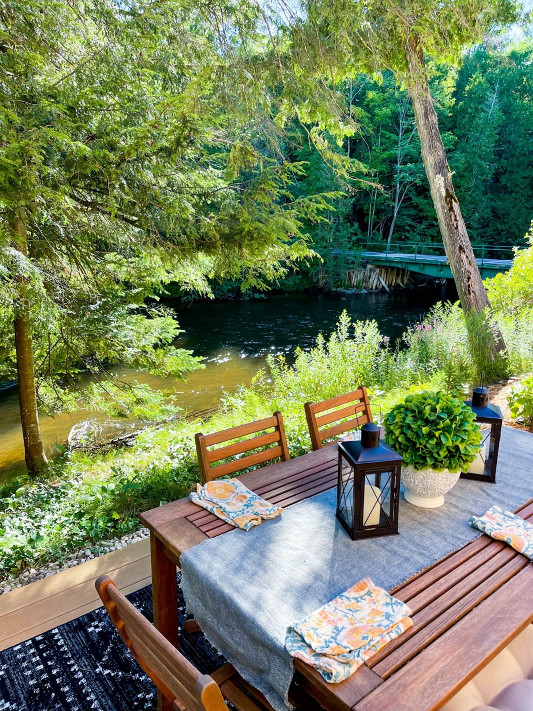 An outdoor dining area with a table set for two, surrounded by lush greenery and a river.