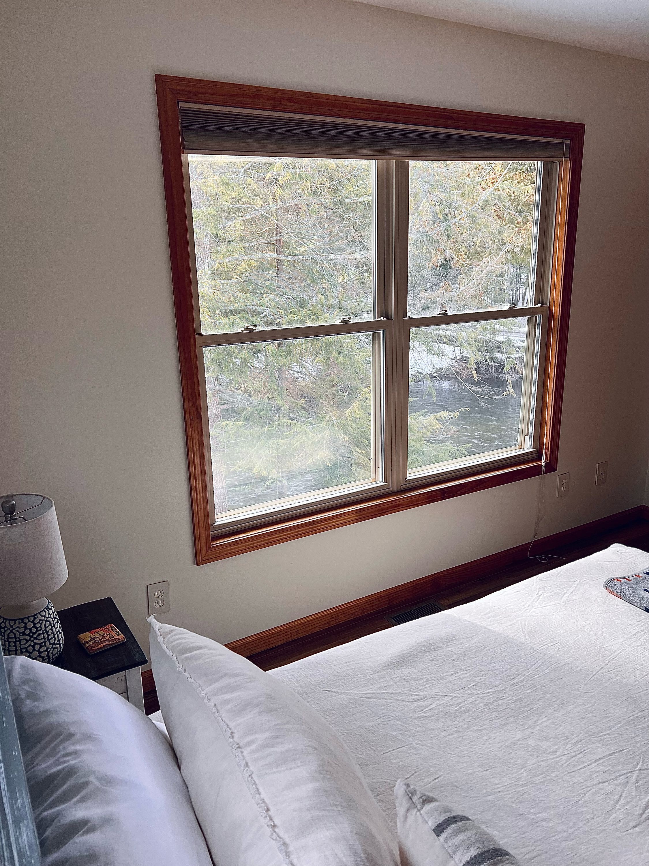 A bedroom with a large window, a bedside table with a lamp, and a bed with white bedding.