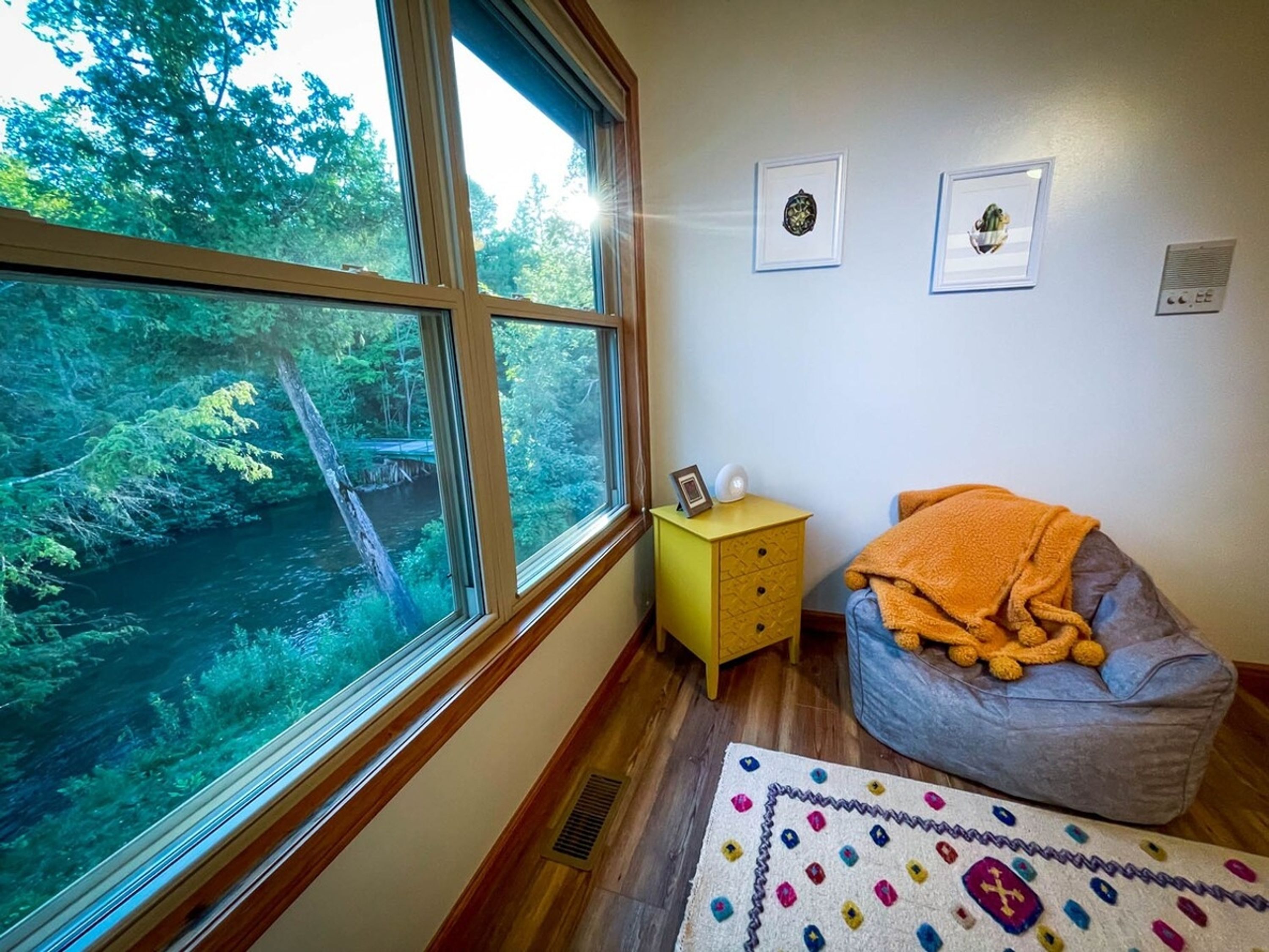 Una habitación con un ventanal grande que da a un bosque, una mesita de noche amarilla pequeña, una silla gris con una manta naranja y una alfombra colorida.