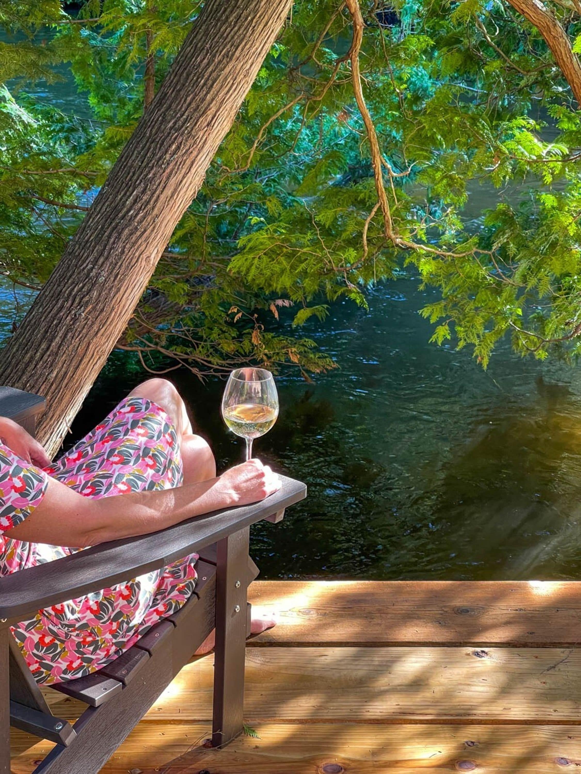 A person sitting on a wooden deck by a body of water, holding a glass of white wine.