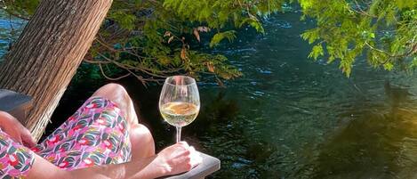 A person sitting on a wooden deck by a body of water, holding a glass of white wine.