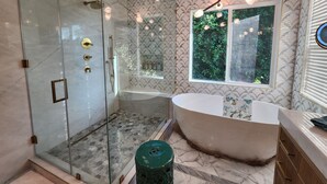 A modern bathroom with a glass-enclosed shower, a freestanding bathtub, and a window with a view of greenery. - Charming 4-bedroom house in lovely Los Angeles with AC (Los Angeles)