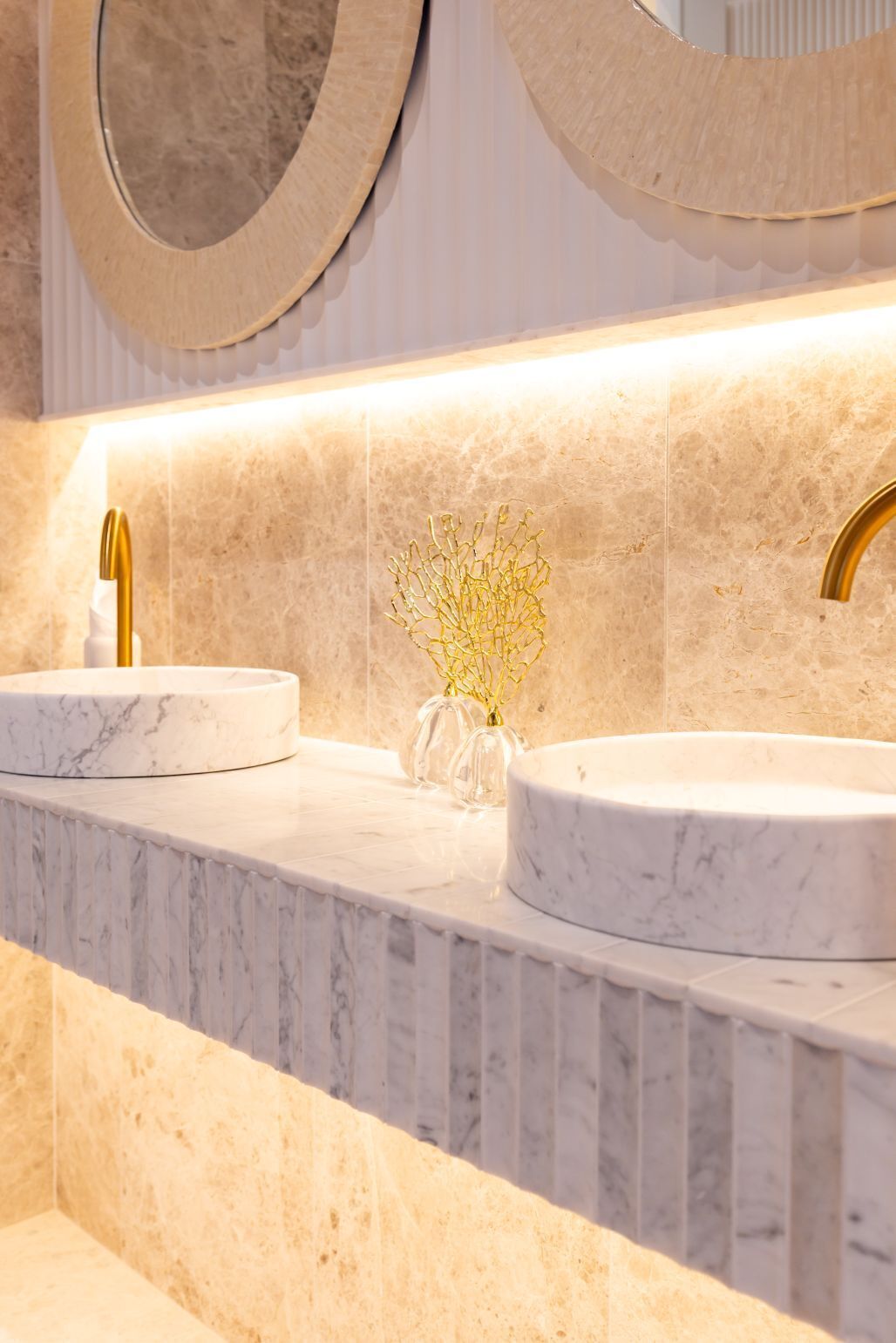 Un baño con dos lavamanos, encimeras de mármol, accesorios dorados y un jarrón decorativo.