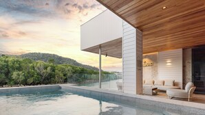 A modern outdoor pool area with a wooden ceiling, a seating area with a sofa and chairs, and a view of a hillside.