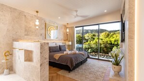 A modern bedroom with a large bed, a TV mounted on the wall, and a view of greenery outside.