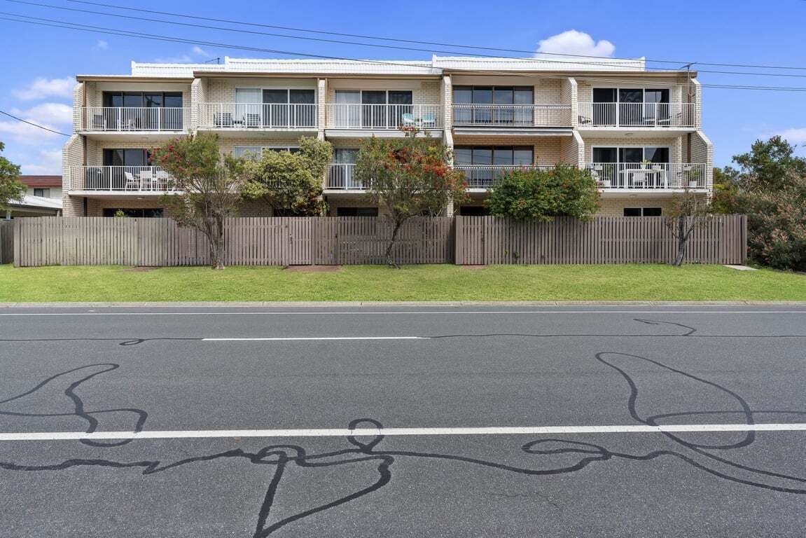 A two-story residential building with a balcony on each floor, a fenced yard, and a clear road in front.