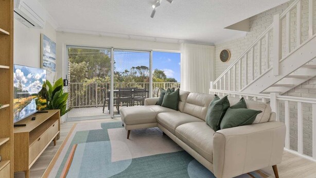 A modern living room with a sectional sofa, a flat-screen TV, and a balcony with a view of trees and a clear sky.