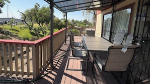 Um pátio externo com mesa e cadeiras, área coberta com teto de vidro e vista para uma paisagem verdejante.