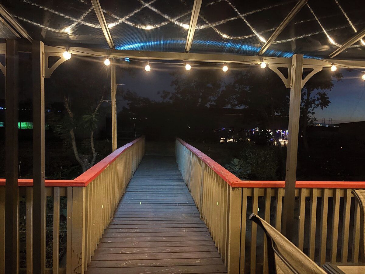 A wooden bridge with railings and string lights, leading to a covered area.