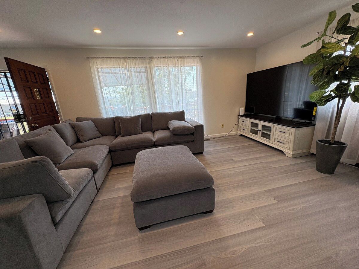 Una sala de estar con sofá seccional, reposapiés, mueble para televisor y planta en maceta.