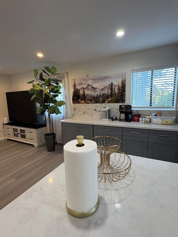 Una cocina moderna con encimera de mármol, un dispensador grande de toallas de papel, una planta en maceta, un televisor de pantalla plana y una ventana con persianas.