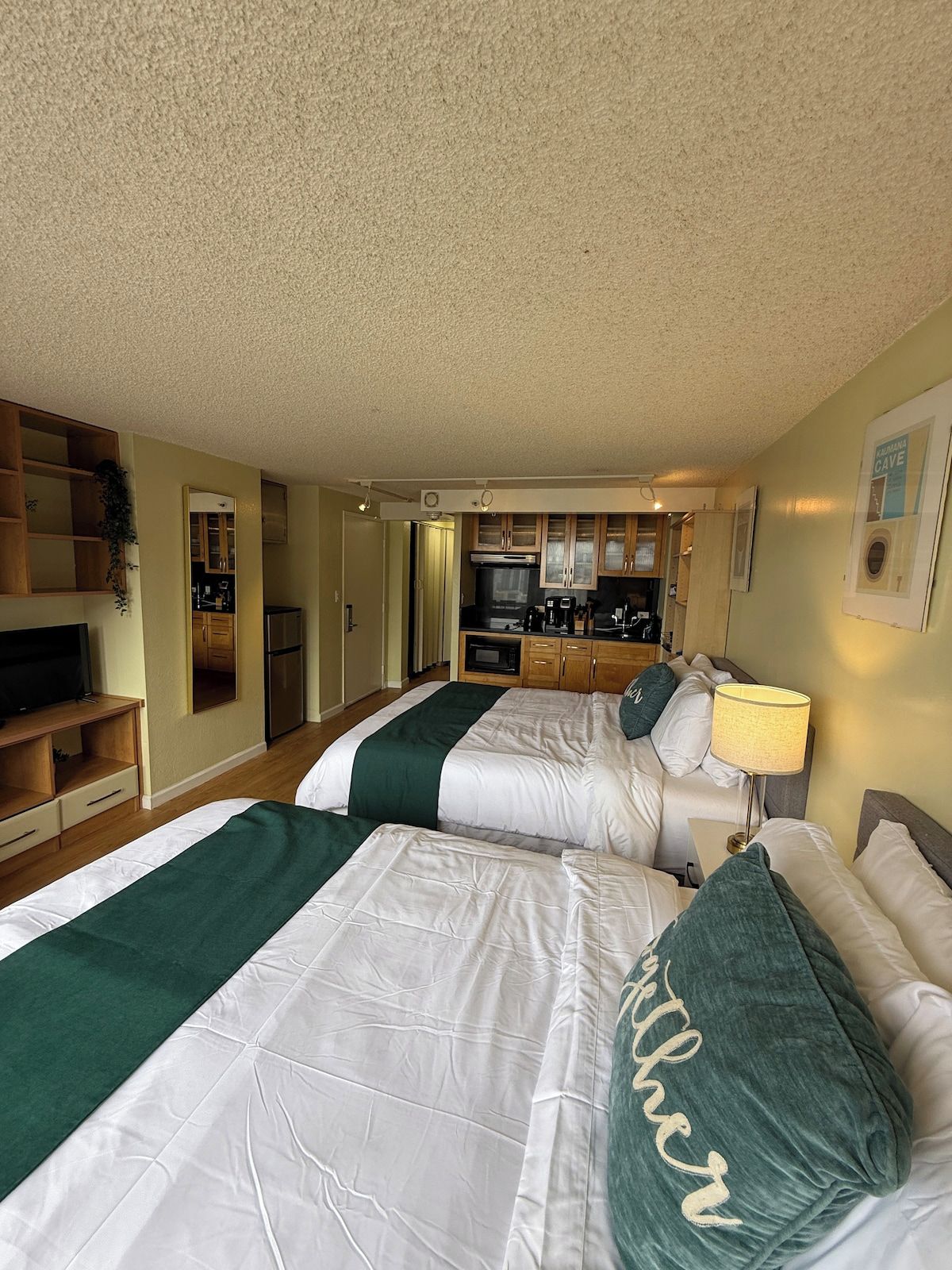 Habitación de hotel con dos camas, kitchenette, televisor y espejo.