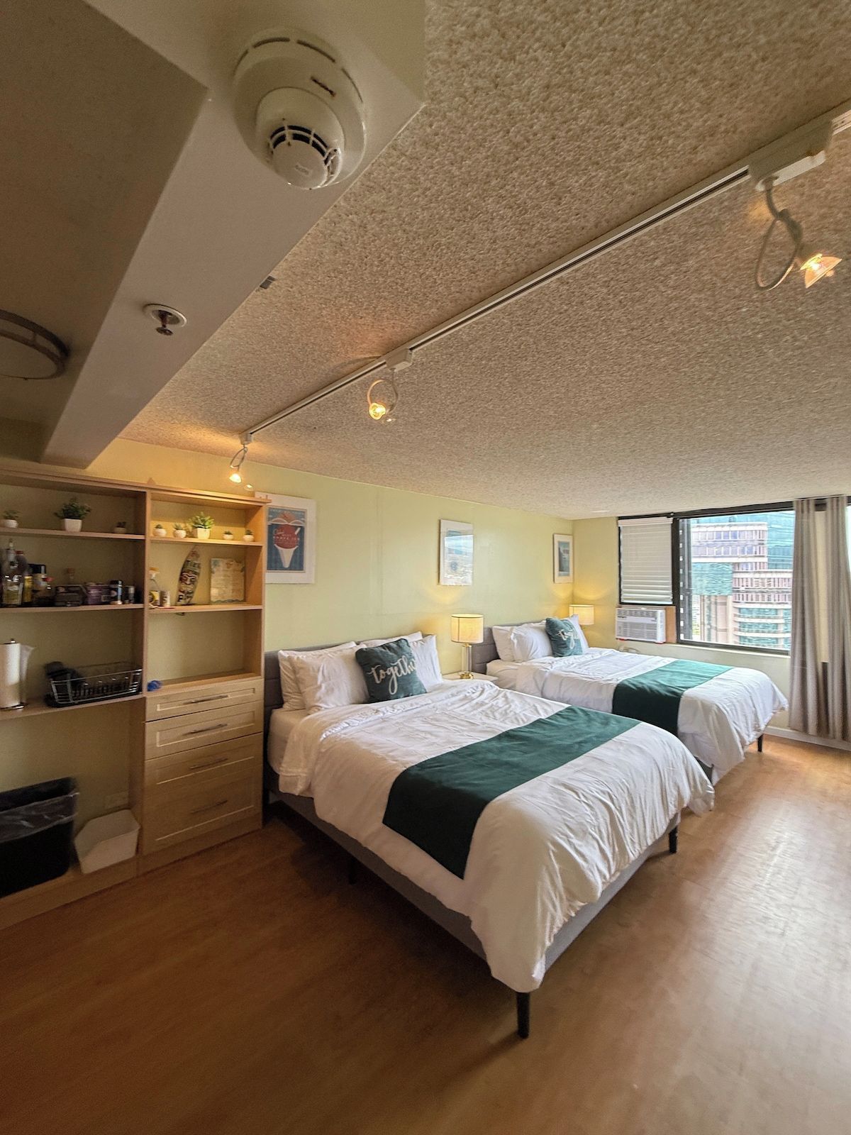 Une chambre d’hôtel avec deux lits, une commode en bois, une étagère avec des articles de toilette et une fenêtre donnant sur la ville.