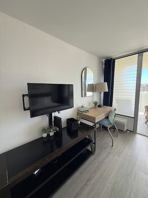 Et moderne hotelværelse med et fladskærms-tv, et skrivebord, en stol, et lille bord med en lampe og et stort vindue med gardiner.