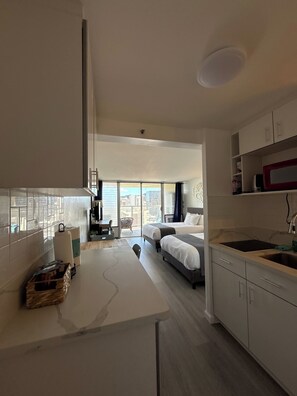 A modern hotel room with a kitchenette, a dining area, and a large window offering a city view.