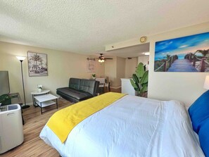 A hotel room with a bed, a sofa, a desk, a ceiling fan, a wall-mounted TV, and a painting.