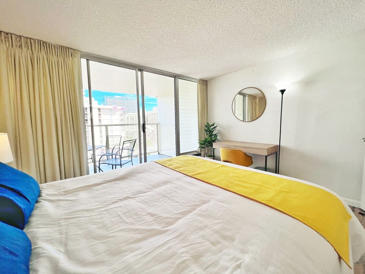 A hotel room with a large bed, a desk, a chair, a potted plant, and a view of the city through the window.