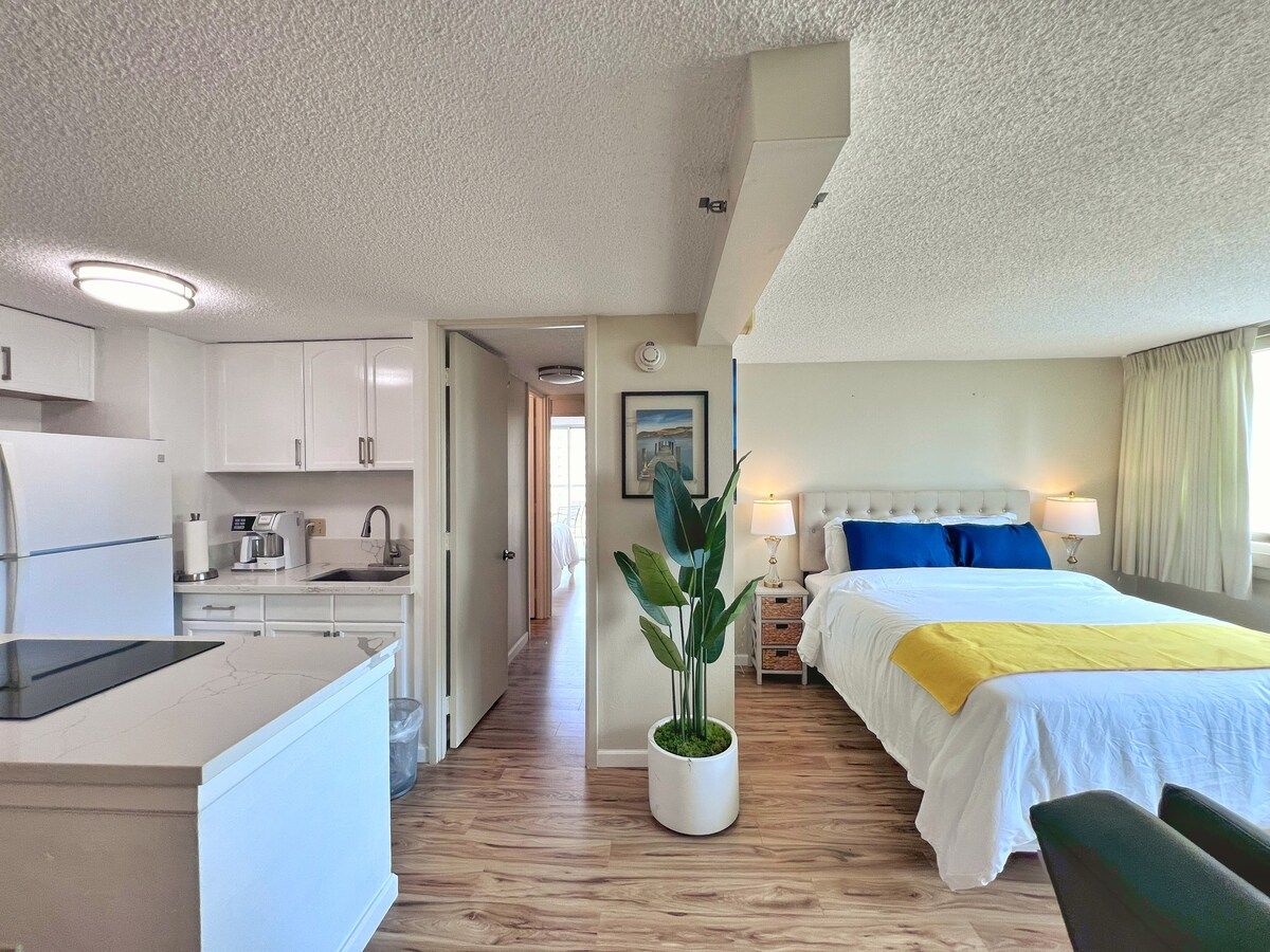 A hotel room with a bed, bedside table, lamp, and a view of the hallway.