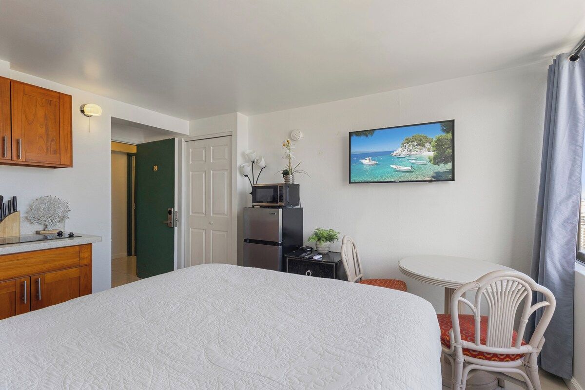 A bedroom with a bed, a desk, a chair, a TV, and a painting of a coastal scene.
