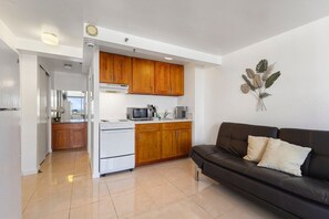 A compact kitchen with wooden cabinets, a white refrigerator, a microwave, and a stove. There is a black leather sofa with cushions in the living area. - Skyline Island Stay • Two Adjoining Studios (Honolulu)