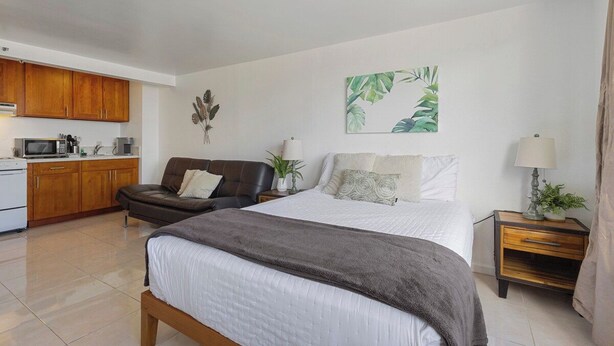 Un dormitorio con cama, mesita de noche, lámpara y vista al área de la cocina.
