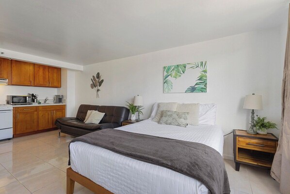 Un dormitorio con cama, mesita de noche, lámpara y vista al área de la cocina.