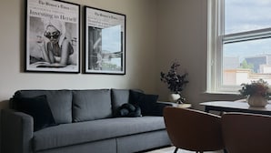 Ett modernt vardagsrum med en grå soffa, svarta kuddar, ett träbord och en kristallkrona. Två inramade tidningsartiklar hänger på väggen.
